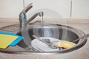 Washing dishes. Forks, spoons, plates of different sizes, rags and a sponge in the sink under the tap