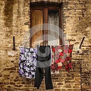 Washing hangs on a line in front of a half shuttered window in a brick wall of a Tuscan village in a square format.