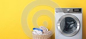 Washing machine with laundry near clean bath towels in wicker basket on yellow wall background