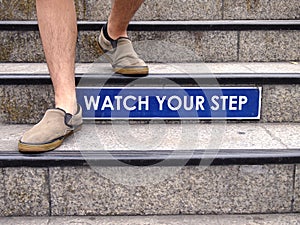 Watch your step sign on a staircase and a pair of feet