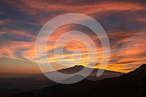 Watching beautiful sunset behind volcano Mount Etna near Castelmola, Taormina, Sicily. Clouds with vibrant red orange colors