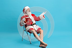 Full-length portrait of senior man wearing Santa Claus costume, holding glass of lager foamy beer isolated over blue