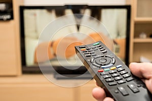 Watching TV and using black modern remote controller. Hand holding TV remote control with a television in the background