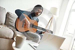 Watching video tutorial. Young man sitting on sofa at home and learning guitar online. Sitting on sofa at home and photo