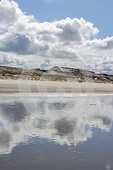 Water on the beach with mirror reflection of white clouds and dunes in the background