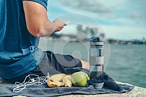 Water bottle, banana, earphone, green apple and towel on the ground beside young man rest after exercise and playing mobile phone