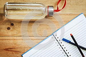 Water bottle, an opened notebook and pens on wooden table background