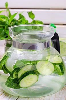 Water with cucumber, mint and ice in glass jugful on wooden table, closeup.