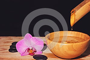 Water drips from the bamboo in a bowl on a wooden table, next to the spa treatment stones and orchid flower, copy space for your t