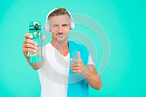 Water is essential for body. Fit athlete in headphones point finger at water bottle on blue background. Drinking water