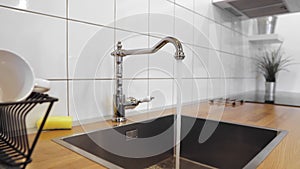 Water flowing out of stainless steel tap. Dish rack holds dishes against wooden countertop, white wall tiles, sink and