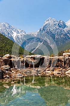 Water is flowing over some rocks of the Blue Moon Valley Lake with the Jade Dragon Snow Mountain in Yunnan, China in the backgroun