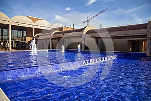 A water fountain in a pool with water falling down steps on the blue tile and words surrounded by buildings and plants and people