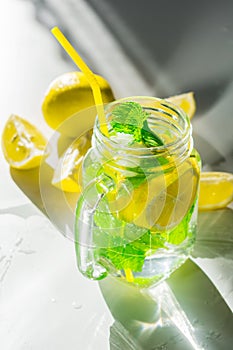 Water with lemon and mint in a transparent mug on a white table, with lemon slices, illuminated by the very bright rays of the sun