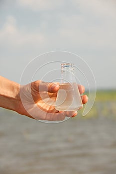Water Purity Test. Hand holding chemical flask with liquid, lake or river in the background.