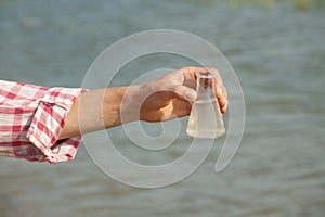 Water Purity Test. Hand holding chemical flask with liquid, lake or river in the background.