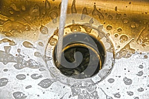 Water running into a garbage disposal of a stainless steel sink with gold toned blurred background and sharp silver foreground