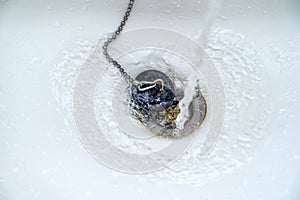 Water running in a white wash basin or bathtub with an old black rubber plug and a rusty drain, copy space, selected focus