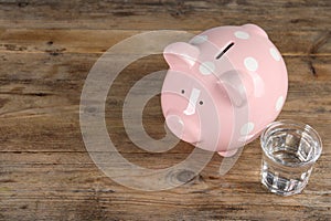 Water scarcity concept. Piggy bank and glass of drink on wooden table, space for text