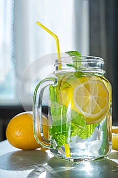 Water with sliced â€‹â€‹lemon and mint leaves in a transparent glass mug with a yellow straw next to the window with morning light