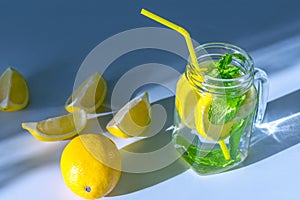 Water with sliced â€‹â€‹lemon and mint leaves in a transparent glass mug with a yellow straw on a table in the morning light