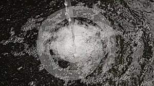 Water stream falls on dark surface, creating air bubbles, drop splashes after falling. Irrigation water flow from pipe