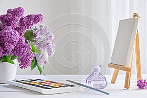 Watercolor paints, canvas on easel and lilac flowers bouquet on table
