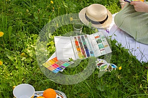 Watercolor palette laying on green grass near woman drawing and painting on plein air in the summer park on sunny day