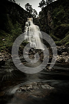 Waterfall cascading down a rocky mountainside surrounded by lush green trees in Scottish Highlands