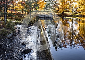 Waterfall on a lake with open floodgates framed by autumn maples lit by the sun in Vermont