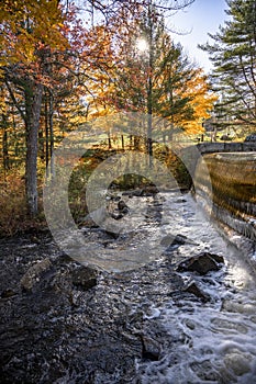 Waterfall on a lake with open sluices framed by autumn maples lit by the sun and reflecting in the water in Vermont