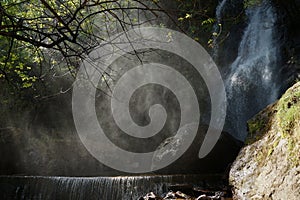 Waterfall and lower cascade in the forest with water fog lit with sunbeams