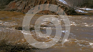 Waterfall on river near mountain at spring with naked bushes, flood, high water.