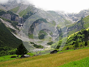 High mountain waterfall rivulets into green valley with huts in alpine terrain