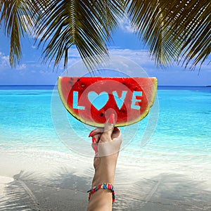 Watermelon slice with text love, woman hands holding it against blue sky and Indian ocean. Summertime concept.Tropical island