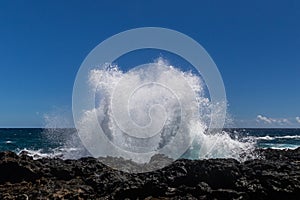 Wave breaking on black lava rock in Hawaii.White spray in the air. Ocean, blue sky and clouds in background.