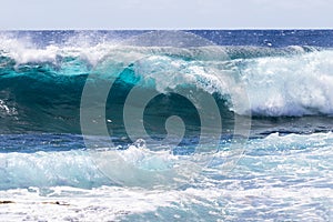 Wave breaking offshore in Hawaii; foam in foreground. Blue ocean, sky, clouds in background.