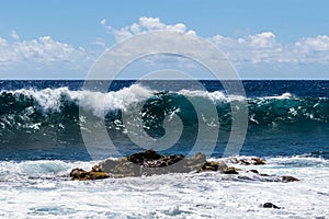Wave breaking offshore in Hawaii; volcanic rock and foam in foreground. Blue ocean, sky, clouds in background.