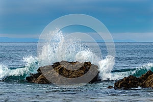 Wave breaking on rock near shore. White spray in the air. Ocean, blue sky, clouds and Catalina island in the distance.