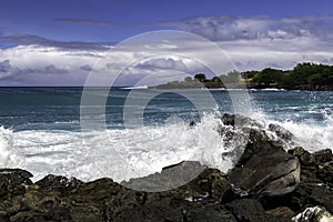 Kona Coast, Hawaii`s Big Island. Wave breaking on rocky shore. Blue-green ocean. Shoreline with trees beyond. Cloudy blue sky.