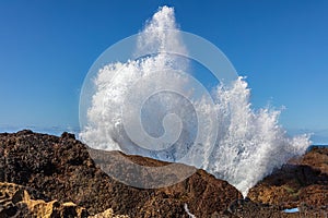 Wave crashing against rocks spray thrown up in the air. Blue sky beyond