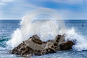 Wave crashing on rock near shoreline. Spray in air; ocean behind; blue sky and clouds above.