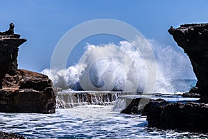 Wave crashing on shore, framed on both sides with rocks. Small waterfall in foreground.