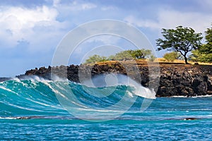 Wave on Hawaiian coast; trees on rocky shore. Clouds in background.