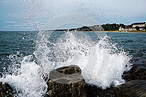 The wave strikes against the stones. Oil painting picture. In the background hill with small houses and trees