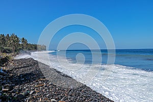 Waves attack the stony beach, and sea water spills over the beach`s edge. Palm trees and tropical vegetation line the coast