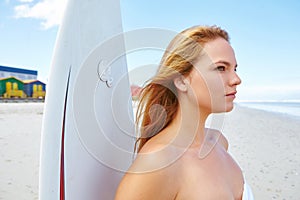 The waves await...a young woman standing on the beach with her surfboard looking out at the ocean.