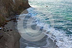 Waves breakaing with foam on sandy beach with backwash against cliff wall