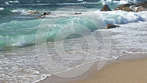 Waves breaking on sandy beach with rocks in background, on a bright sunny day