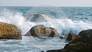 Waves crash against rocks at beach, sea spray in air, sunlight reflects on water at dusk. Natural ocean wave motion, hit
